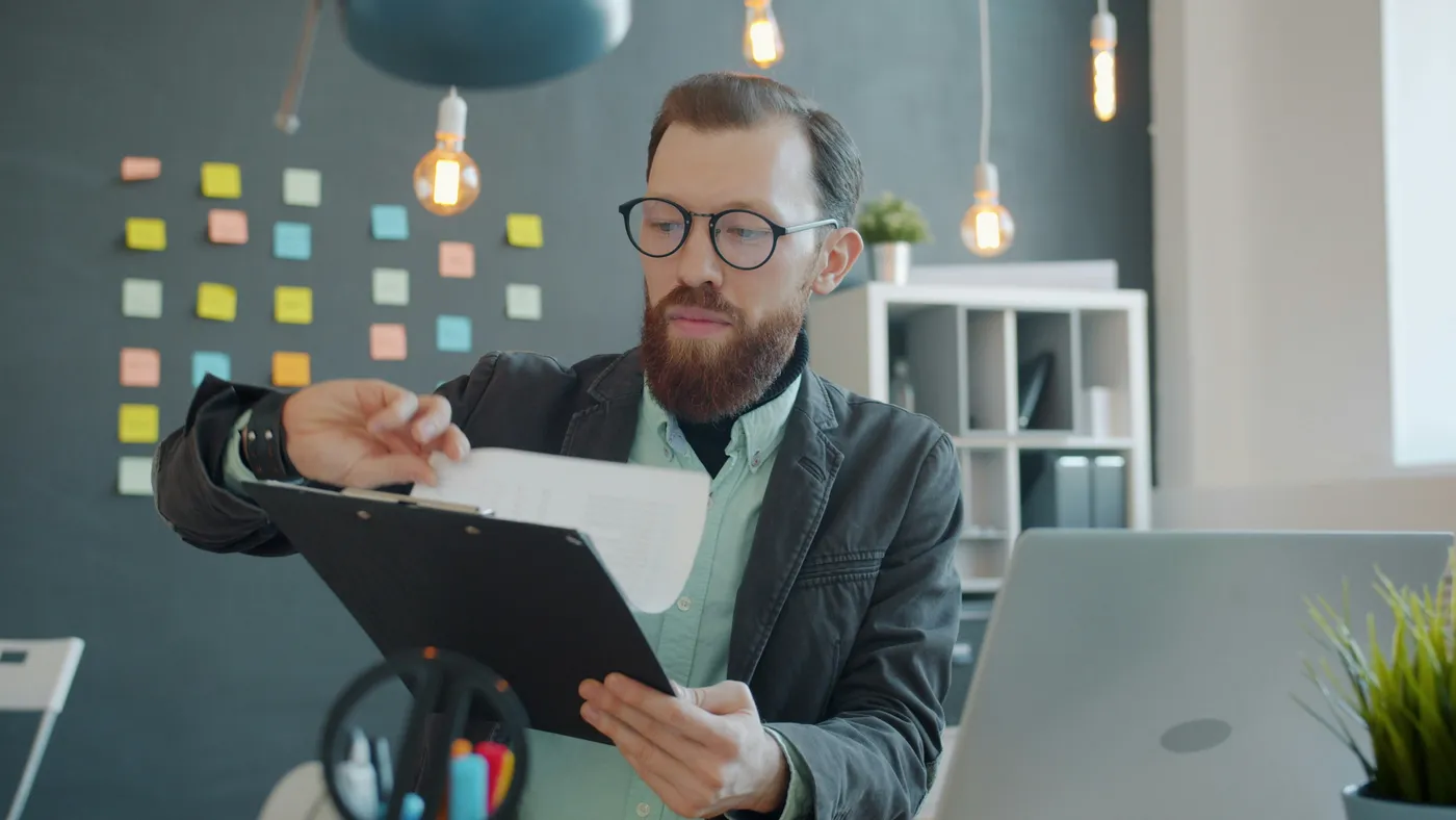 A man at his desk reading a page from a clipboard, a laptop open beside him