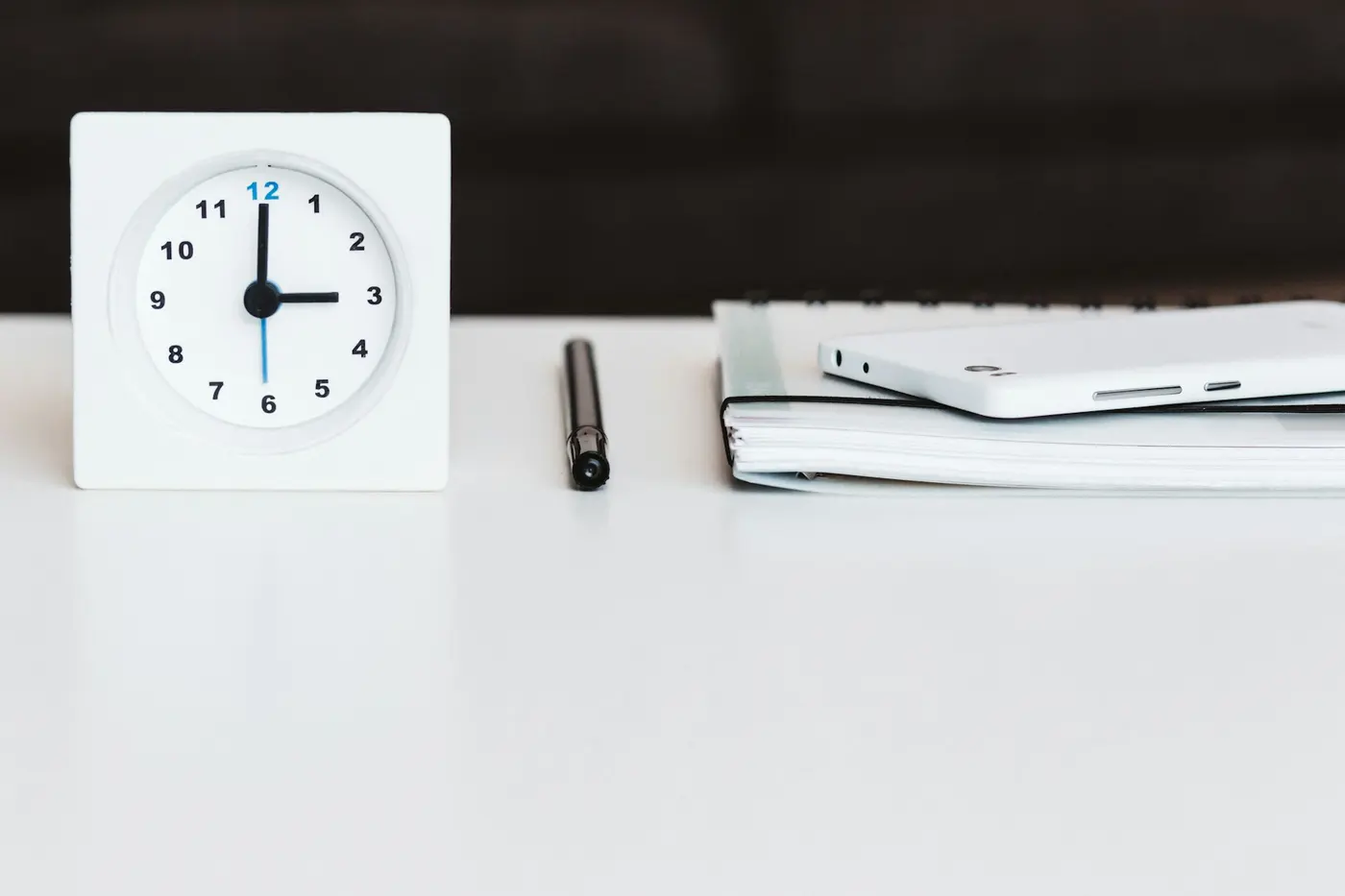 A white desk clock beside a pen and a closed notebook on a white desk