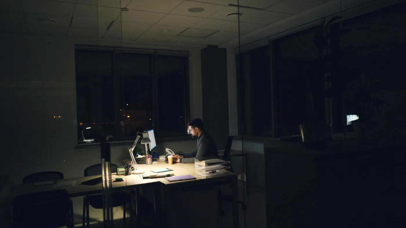 One person working late at a single lit desk in an otherwise dark office