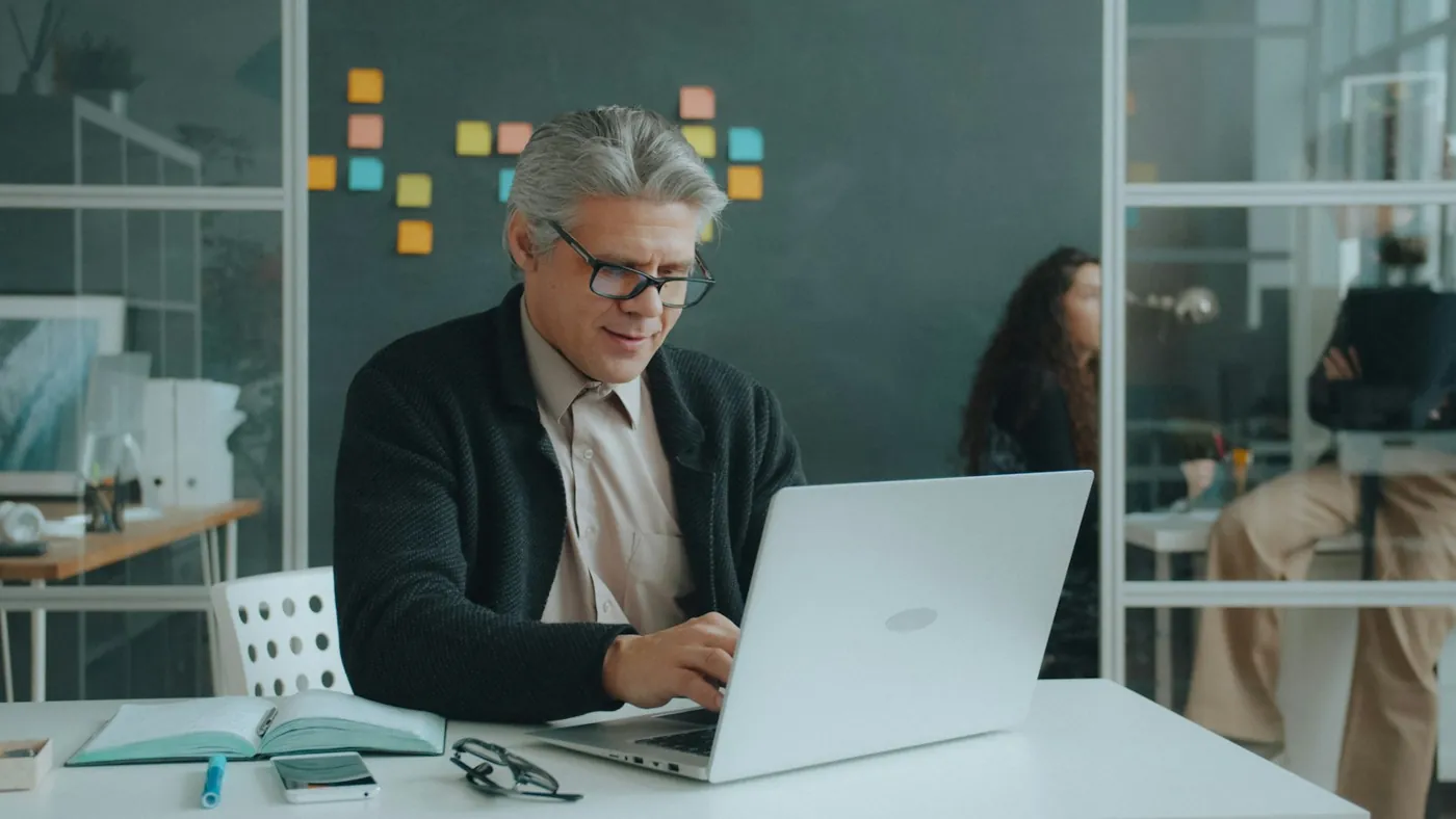 A managing partner working on a laptop in a glass-walled office while colleagues work behind him